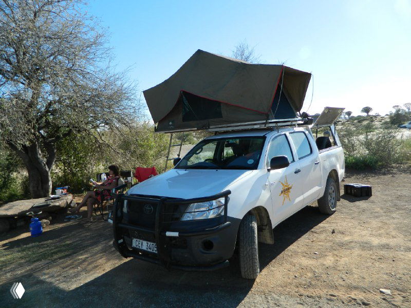 Внедорожник, оборудованный для кемпинга, с раскладывающейся палаткой на крыше, кухонной зоной и вещами у стоянки под деревьями в лагере.