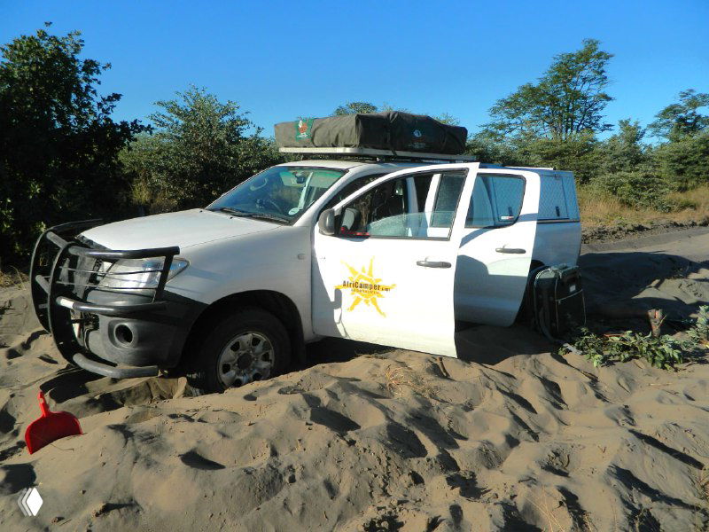 Внедорожник, застрявший в мягком песке, рядом видны следы и попытки вытащить машину — типичная проблема при поездках по дюнам и рыхлым путям.