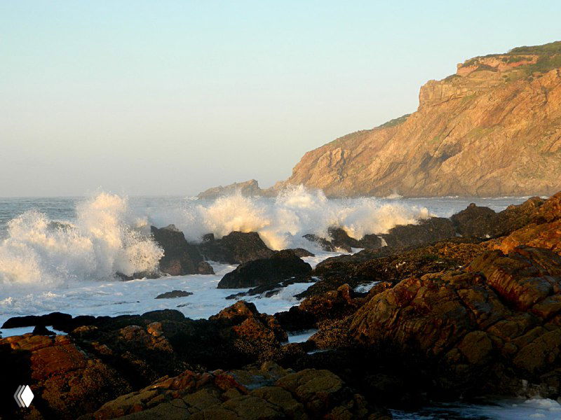 Волны разбиваются о скалистый берег: пенящаяся вода, мокрые камни и крутой прибрежный утёс под дневным светом.