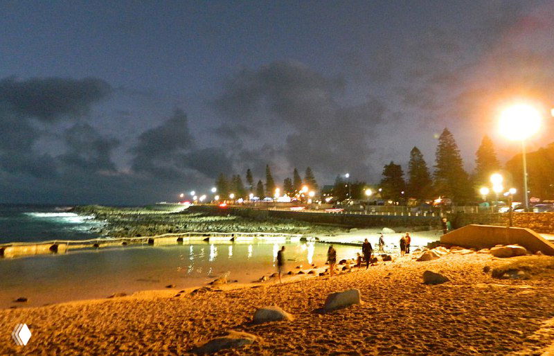 Ночная набережная с освещённым пляжем и людьми: фонари вдоль берега, тёплый городской свет и отражения на влажном песке.