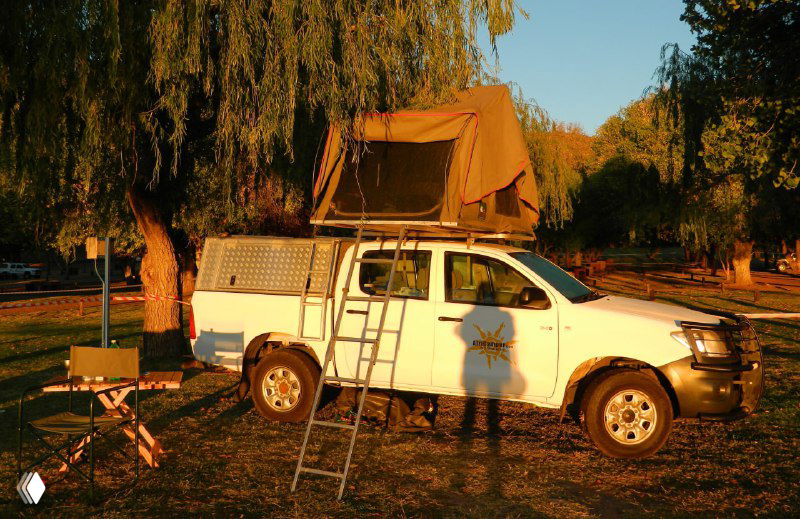 Белый внедорожник с палаткой и лесенкой у кемпинга на фоне зелёных деревьев в золотом свете вечера — типичный автокемпер в ЮАР.