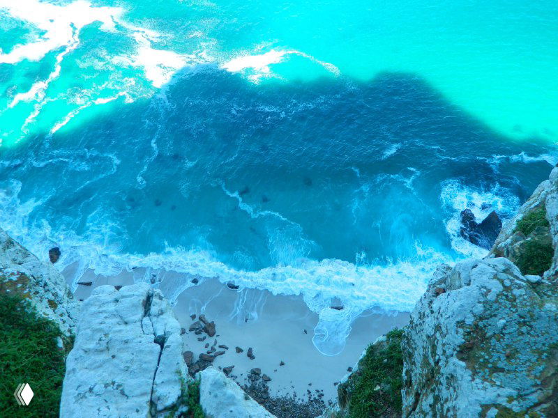 Вид сверху на бирюзовую воду и белый песчаный пляж внизу, волны накатывают у подножия высокийх известняковых скал.