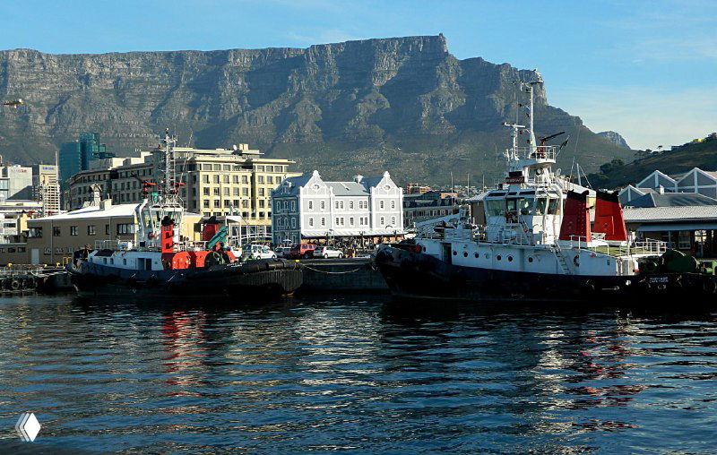 Причал и старые голландские домики у воды, яхты и небольшие рабочие суда на фоне массивной Столовой горы под голубым небом.