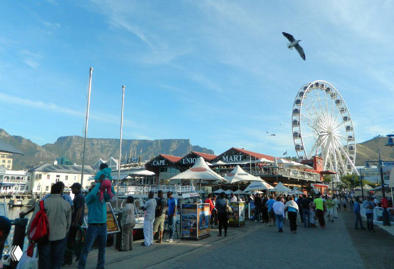 Народная суета на Ватерфронте: толпа людей, прибрежные кафе и большое колесо обозрения на фоне Столовой горы и побережья.