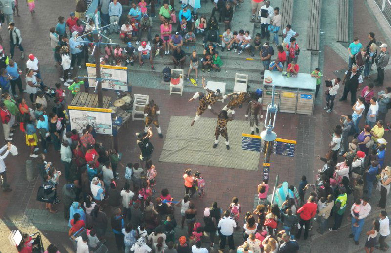 Уличное представление на Ватерфронте: большая толпа вокруг уличных артистов, вид сверху на людей и оживлённую прогулочную зону.