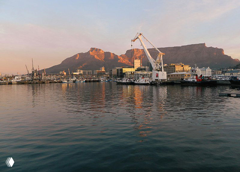 Рассветный или закатный вид на порт и кран, тёплые тона света отражаются в воде, на заднем плане контуры Столовой горы и городских сооружений.