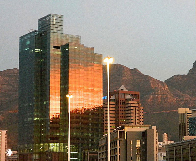 Современные стеклянные офисные здания в городе, отражающие свет заката на фоне силуэта Столовой горы и городского горизонта.