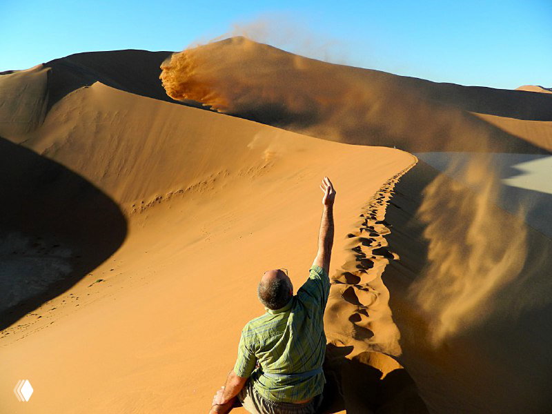 Человек на вершине дюны подбрасывает песок рукой; видны крутые скаты, следы шагов и игра света на песке.