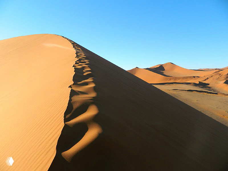 Гребень дюны с четким следом от шагов на вершине и резким контрастом теней и света под ясным голубым небом.
