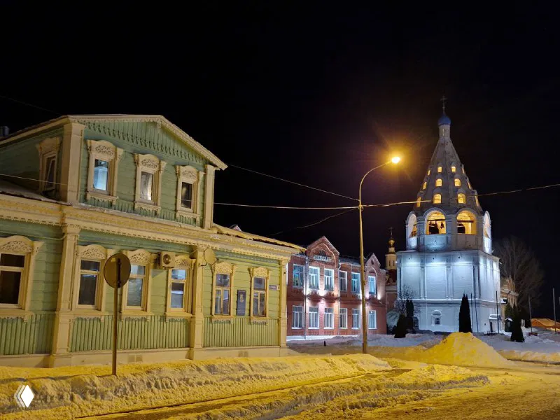 Ночная улица с деревянными домами и освещённой церковной башней, историческая застройка и заснеженный тротуар под уличными фонарями.