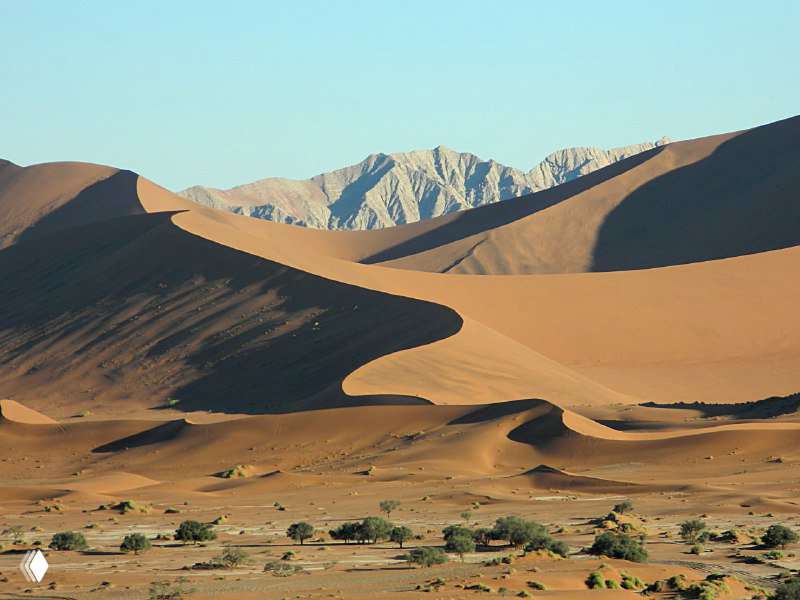 Широкий план долины с волнистыми барханами и дуновением света; на дальнем плане видны горы за песчаными склонами.