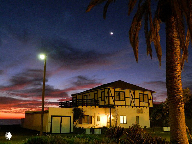 Ночной пейзаж с фахверковым домом у океана, тёмное небо, пальма на переднем плане и подсвеченное здание на фоне сумерек.