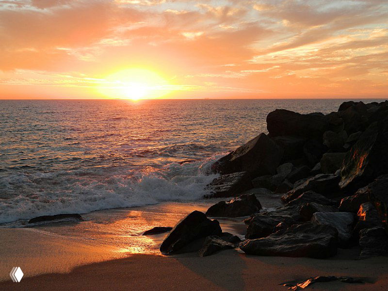 Закат над океаном: солнце опускается за горизонт, золотистые блики на волнах и скалистый берег в тёплых тонах.