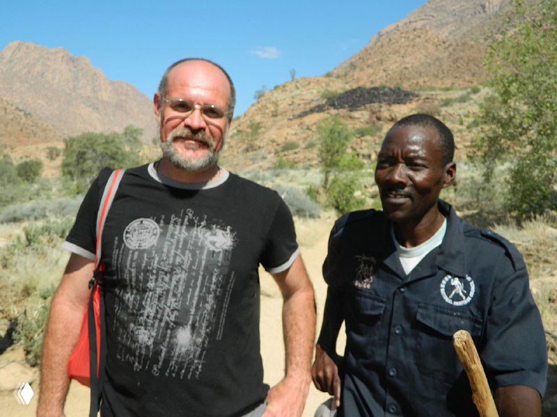 Два человека у реки на фоне гор — сопровождающий гид из народа дамара и посетитель; видны трость гида и горный фон, без указания личности.