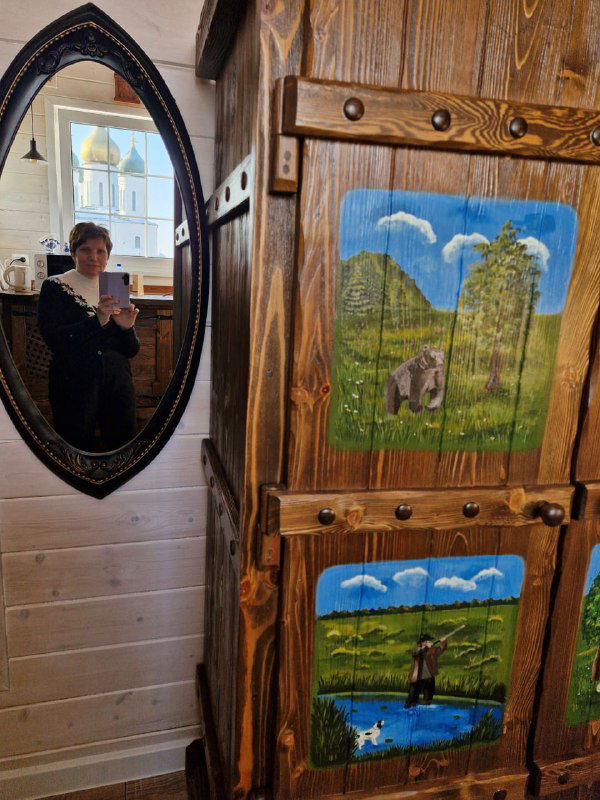 Интерьер гостевого дома: зеркало с отражением окна и церкви, рядом расписной деревянный шкаф и элементы деревенского декора.
