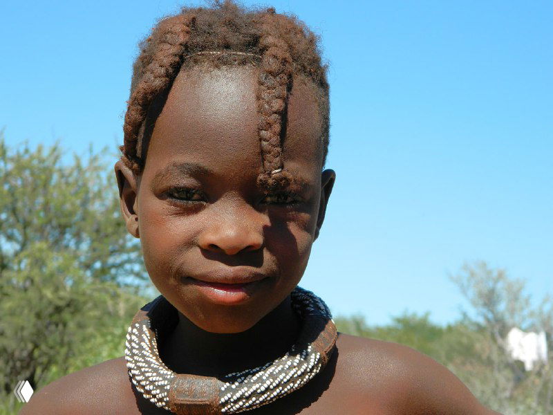 Портрет девочки химба с короткими косичками и плотным ожерельем, светлый небесный фон и тонкая растительность вокруг поселения.