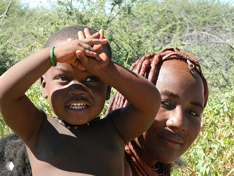 Женщина химба держит на руках ребенка: солнечный свет, близкий план лиц, животные и растительность на заднем плане дают контекст деревни.