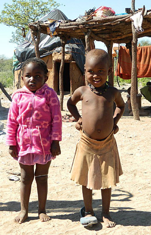 Двое маленьких детей химба стоят у простой конструкции хижины, на одном ребёнке розовая кофточка, на другом — традиционная юбочка и босые ноги.