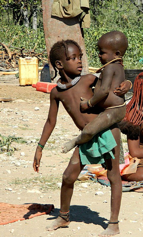 Дети играют и носят друг друга на руках в деревне химба: коричневые тона кожи, простая одежда и фон из кустарника и хозяйственных предметов.
