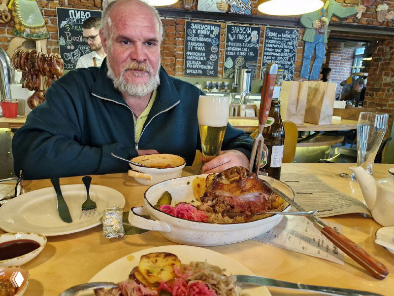 Тарелка с запечённым мясом и гарниром на столе пивного бистро; рядом бокал пива и довольный посетитель в колоритной обстановке.