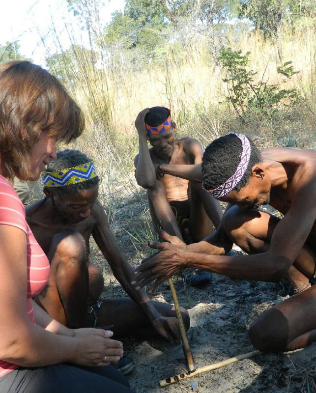 Сцена в деревне сан: несколько человек сидят в кругу на песке и демонстрируют технику добычи огня трением палочек, туристка наблюдает рядом.