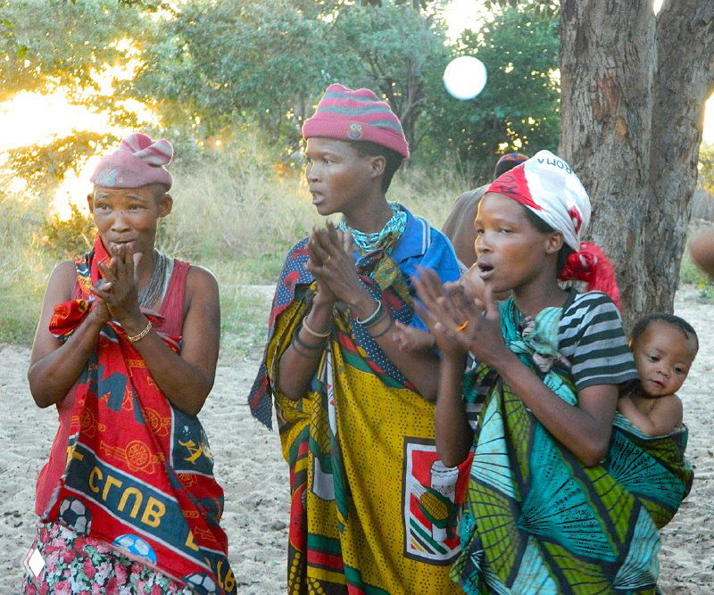 Женщины племени сан в традиционных цветных одеждах поют и хлопают в ладоши, одна женщина с ребёнком на спине, вечерний ансамбль общинного танца.