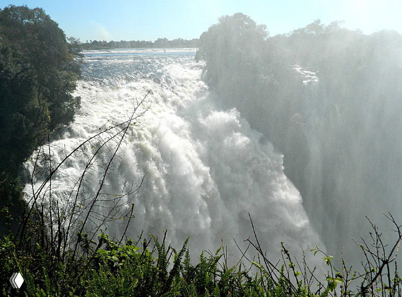 Мощный поток воды, падающий с уступа водопада Виктория, белая пена и столб брызг на фоне густой прибрежной растительности.