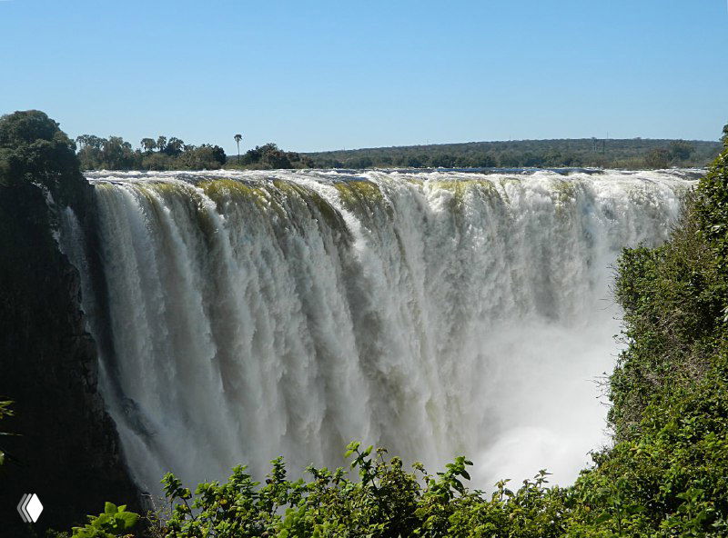 Широкий вид на подковаобразную стену водопада Виктория, каскады воды и облако брызг, обрамлённое зеленью на переднем плане.