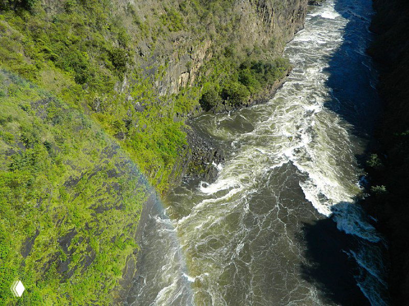 Вид сверху на бурную реку в каньоне Замбези: быстрые потоки, скалистые берега и дуга радуги, возникающая из брызг водопада.