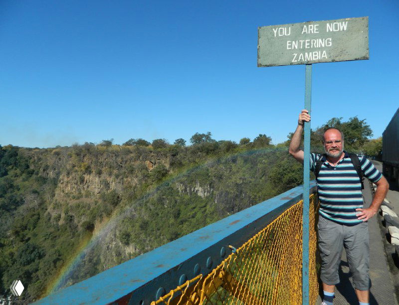 Турист стоит у знака 'YOU ARE NOW ENTERING ZAMBIA' на мосту — отметка границы, видна ограждённая дорожка и обрывистые склоны.