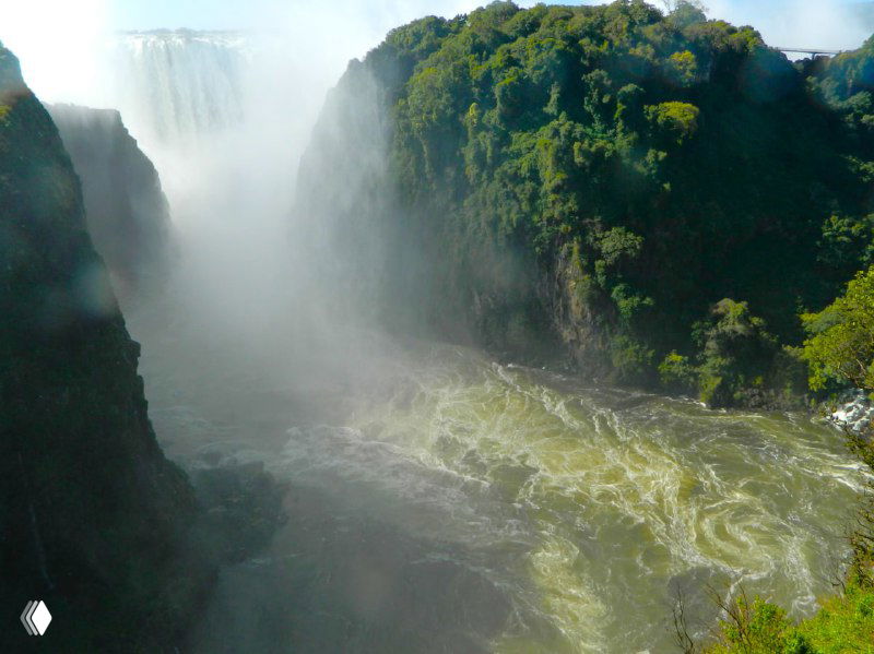 Мощные столбы водяной пыли у подножия водопада Виктория, густой туман от брызг и зелёные обрамления каньона на заднем плане.