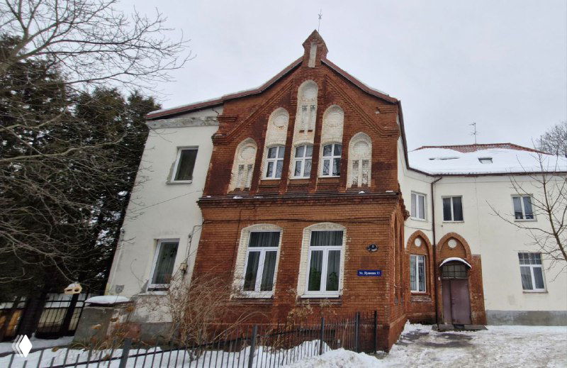 Ещё одна краснокирпичная вилла с декоративным фронтоном и высокими окнами; перед домом кустарники и заброшенные ограждения.