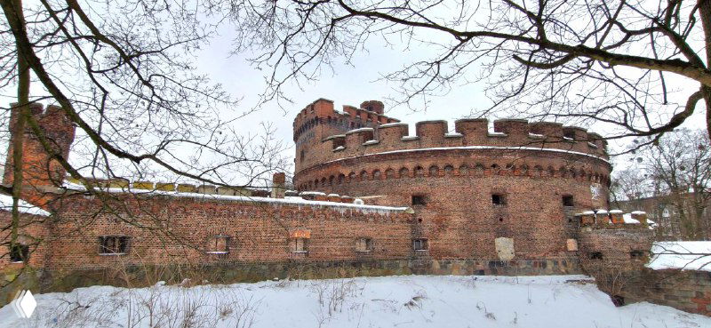 Круглая мощная фортификационная башня из кирпича с зубчатым венцом и земляным основанием на заснеженном берегу.