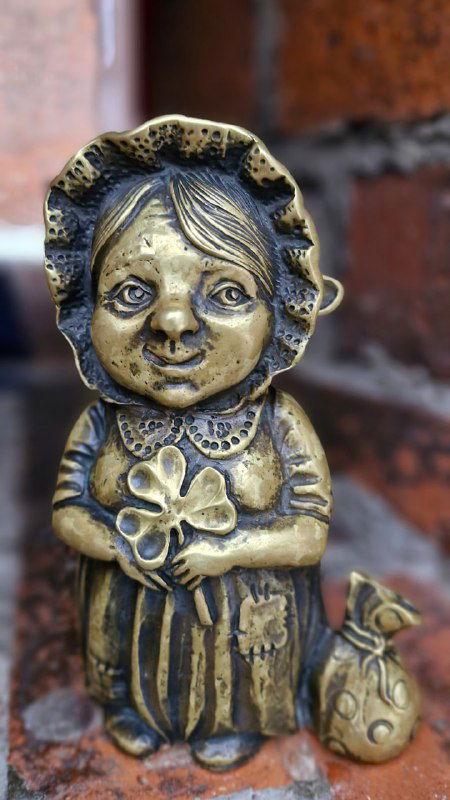 Крупный план бронзовой фигурки бабушки-хомлины с мешочком янтаря у кирпичной стены, видны детали одежды и улыбка скульптуры.