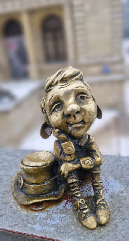 Фигурка папы-хомлина на перилах лестницы рядом с городским зданием, видно мелкую детализацию и архитектурный фон.