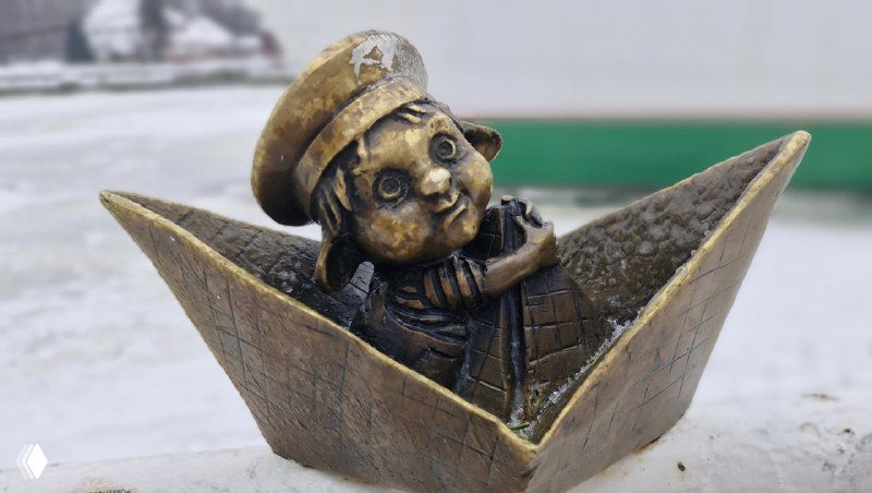 Бронзовый хомлин в бумажном кораблике на набережной, на заднем плане размыт корпус Музея Мирового океана и зимний пейзаж.