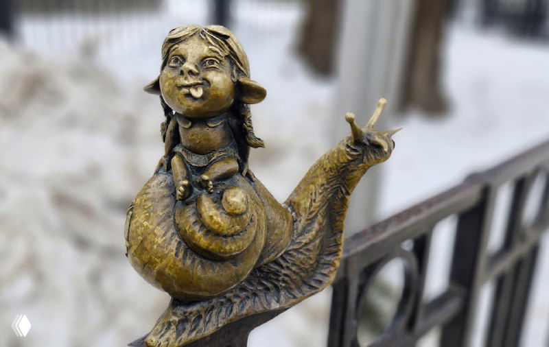 Фигурка хомлинки, едущей на улитке у ограждения — крупный план металлической скульптуры с видимой текстурой и следами снега.