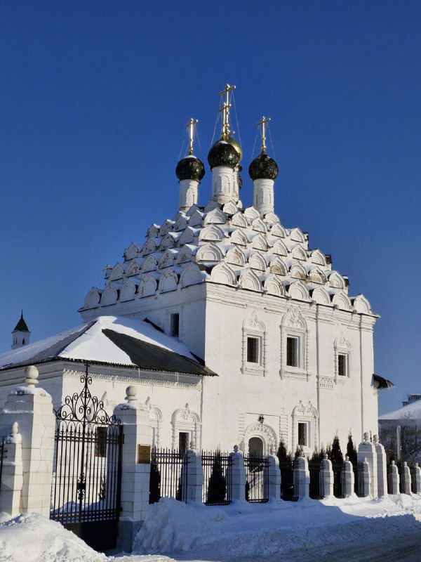 Белая старинная церковь с множеством мелких куполов и узорным боковым фасадом, покрытая снегом.