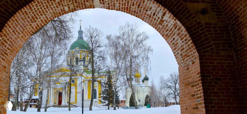 Вид на православный храм через арку кирпичной стены кремля: белые и зелёные купола на фоне зимнего неба и голых деревьев.