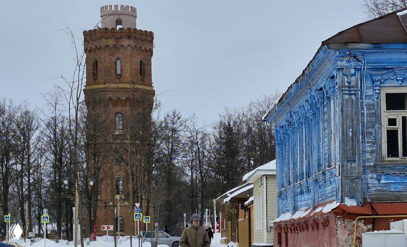 Старая водонапорная башня в районе города и яркий резной синий деревянный дом на переднем плане, зимний городской мотив.
