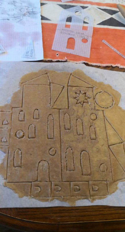 Раскатанное на пергаменте тесто с наложенным бумажным шаблоном: видны контуры дома, линии, разметка и подготовка к вырезке.
