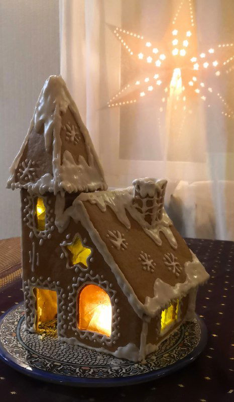Собранный пряничный домик с белой глазурью и внутренней подсветкой от маленькой гирлянды, стоит на блюде у окна со светящейся звездой.