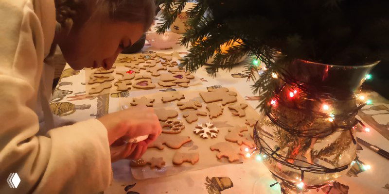 Человек украшает множество печенек и пряников у рождественской ёлки, аккуратно нанося глазурь и расставляя украшения на столе.