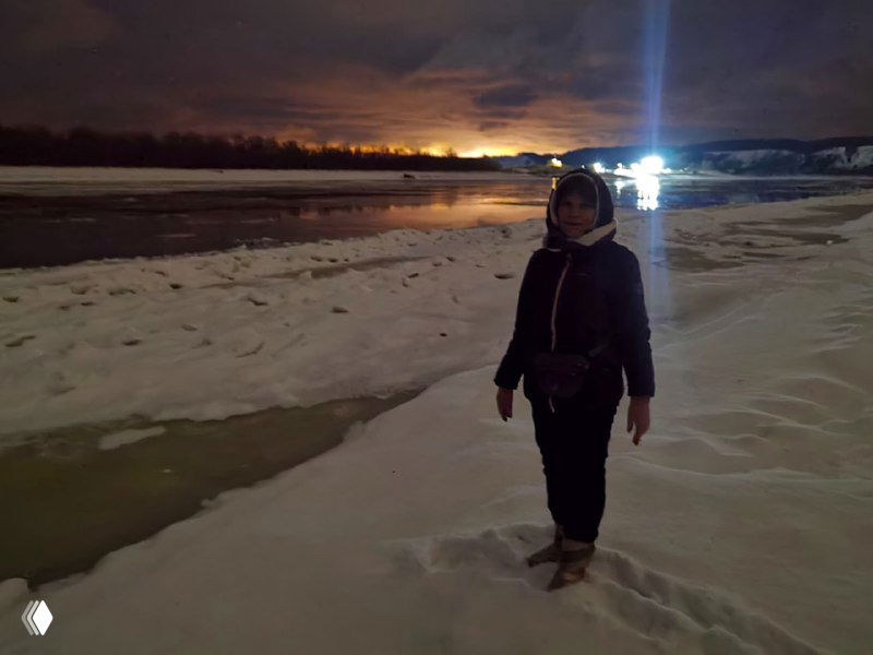 Человек идёт по заснеженному берегу Иртыша вечером; тёмное небо, отражения огней на льду и фигура на переднем плане создают атмосферу прогулки