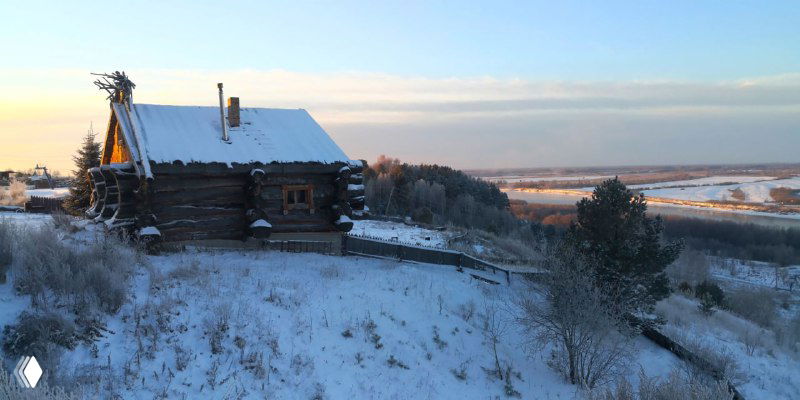Бревенчатый домик на засневшей возвышенности с видом на реку и просторный зимний пейзаж вокруг — типичная усадьба в туристическом комплексе