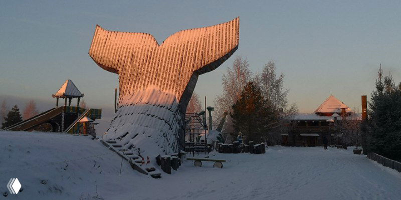 Огромная деревянная фигура хвоста кита на открытой площадке комплекса «Абалак», покрытая снегом, рядом видны деревянные игровые элементы