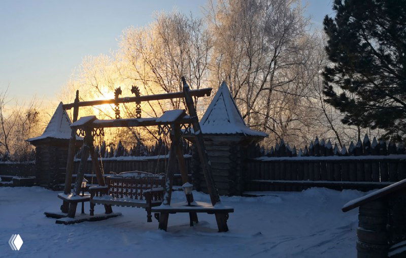 Деревянные качели и декоративные конструкции на заснеженной площади городка «Абалак», тёплый низкий свет рассвета или заката на заднем плане