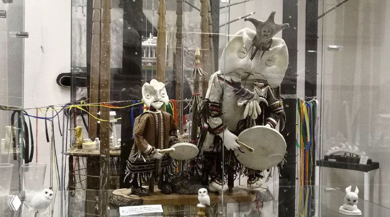 Экспонаты в стеклянной витрине: куклы и фигурки в национальных костюмах, музыкальные инструменты и декоративные детали на фоне музейной инсталляции.