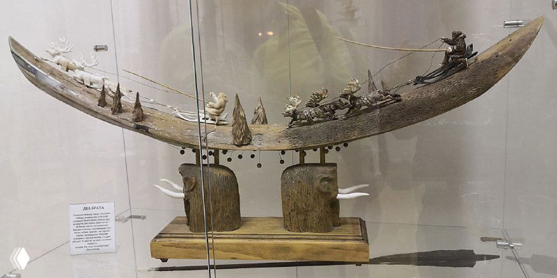 Панорамная композиция на фрагменте бивня мамонта: сценка с фигурками, миниатюрными деревьями и персонажами, выставочная подставка и поясняющая табличка.