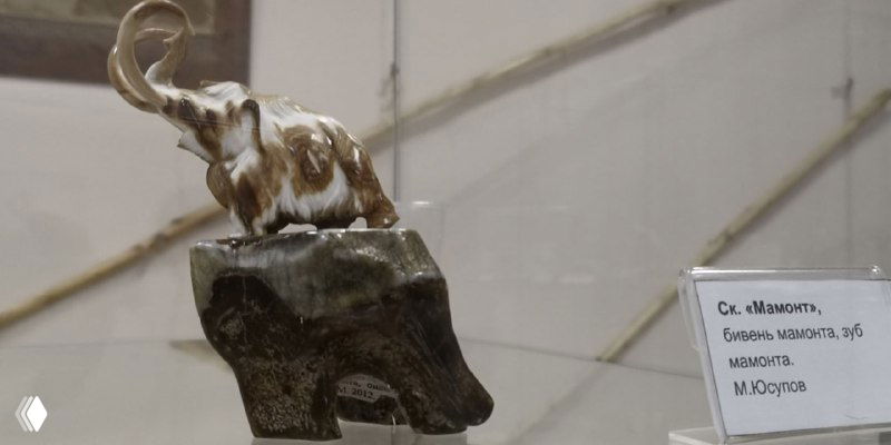 Небольшая скульптура слонёнка, выполненная из кости или бивня мамонта, выставленная на тёмной подставке — пример филигранной работы современных мастеров.